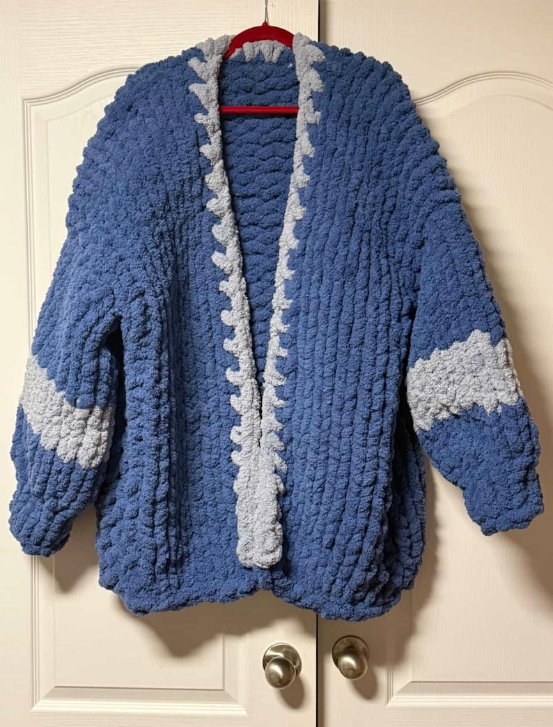 May include: A chunky knit cardigan made from thick blue yarn with a contrasting light gray trim along the open front edges. The sleeves feature a wide horizontal light gray stripe, and the entire garment has a textured, looped appearance.