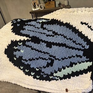 May include: A chunky knit blanket featuring a large central butterfly motif. The butterfly wings are crafted in shades of black, light blue, and mint green, set against a solid white background. The blanket has a thick, textured yarn construction.
