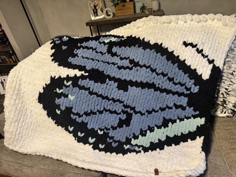 May include: A chunky knit blanket featuring a large central butterfly motif. The butterfly wings are crafted in shades of black, light blue, and mint green, set against a solid white background. The blanket has a thick, textured yarn construction.