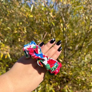 May include: A colorful floral patterned scrunchie with red, blue, green, and white flowers. The scrunchie is tied in a bow and is being held by a hand with black nail polish.