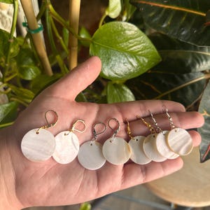 Peut inclure: Une collection de boucles d'oreilles rondes en coquillage blanc, avec différents styles de crochets en métal, or et argent. Les boucles d'oreilles sont présentées sur un fond de feuillage vert.