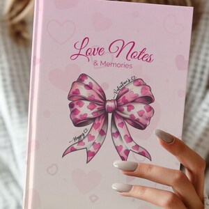 Puede incluir: Una libreta rosa con un lazo rosa y corazones en la portada. El texto en la portada dice "Love Notes & Memories" y "Valentine's".