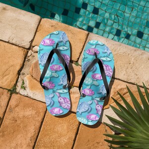 Könnte beinhalten: Zwei Flip-Flops mit einem blauen Aquamuster, das Fische und Schildkröten in Lila-, Rosa- und Blautönen zeigt. Die Riemen sind schwarz. Die Schuhe liegen auf einer Steinoberfläche in der Nähe eines Schwimmbeckens.