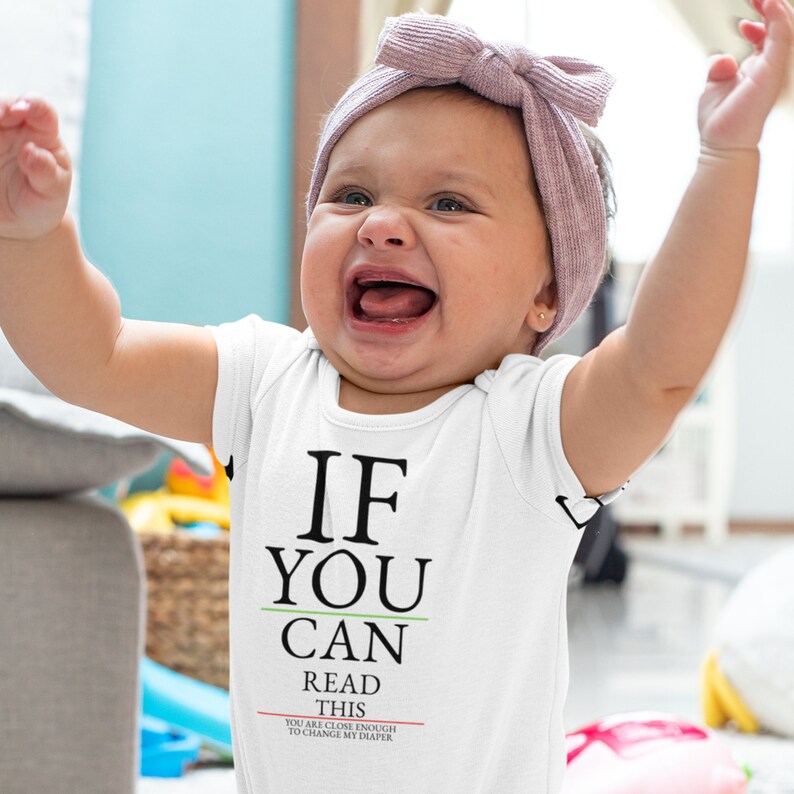 May include: A white baby onesie with a black and green eye chart that reads "If you can read this you are close enough to change my diaper". A baby girl is wearing the onesie and is laughing with her arms raised.