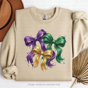 May include: Beige sweatshirt featuring three watercolor bows in purple, green, and gold. The bows have a textured, painted appearance with paint splatter details. The text "Design shown as example only" is at the bottom.