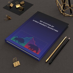 Może przedstawiać: Notatnik z napisem "MY SYSTEM OF STOCK MARKET INVESTING" z niebieską okładką z grafiką byka i niedźwiedzia. Widoczne są również złote i czarne ołówki oraz złote spinacze do papieru.