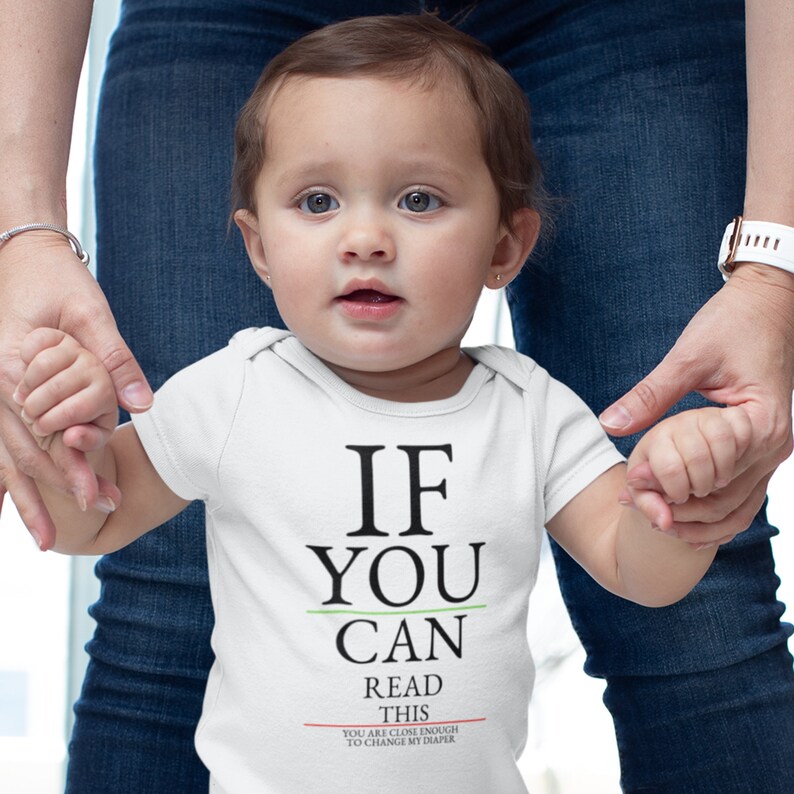 May include: A white baby grow with the text "If you can read this you are close enough to change my diaper". A baby is wearing the grow and holding hands with an adult.