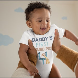 May include: A white baby grow with the text "Daddy's First Round Pick" and a football graphic. The baby is wearing the grow and smiling.