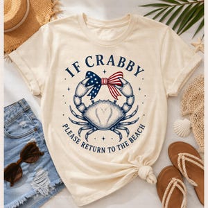 May include: Cream-colored t-shirt featuring a blue crab with a patriotic bow and the text "IF CRABBY PLEASE RETURN TO THE BEACH". The design is in navy blue and red.