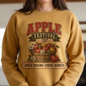 May include: A mustard yellow crewneck sweatshirt featuring a graphic print that reads APPLE FESTIVAL APPLE PASS with an illustration of a wooden basket filled with red and yellow apples, white blossoms, and the text APPLE PICKING CIDER DONUTS.