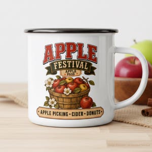 May include: A white enamel mug with a black rim featuring a graphic of a wooden basket filled with red and yellow apples and white blossoms. The text reads APPLE FESTIVAL, APPLE PASS, and APPLE PICKING - CIDER - DONUTS in red and black lettering.