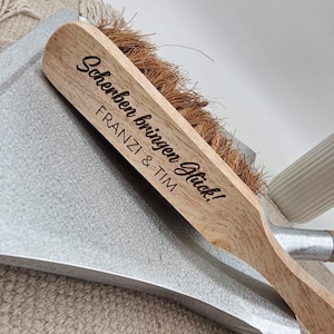 May include: A wooden brush with natural bristles and a metal dustpan. The brush handle is engraved with the words "Scherben bringen Gl&uuml;ck!" and the names "FRANZI & TIM". The dustpan is silver.