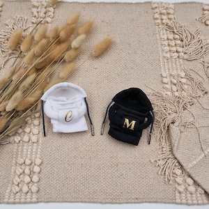 Könnte beinhalten: Zwei Kapuzenpullover mit gestickten Buchstaben. Der weiße Pullover hat ein goldenes "C" und der schwarze Pullover ein goldenes "M".