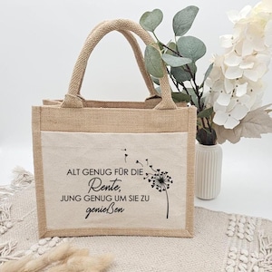 Beige Jutetasche mit schwarzem Aufdruck einer Pusteblume und dem Spruch „Alt genug für die Rente, jung genug um sie zu genießen“, dekoriert mit Trockenblumen.