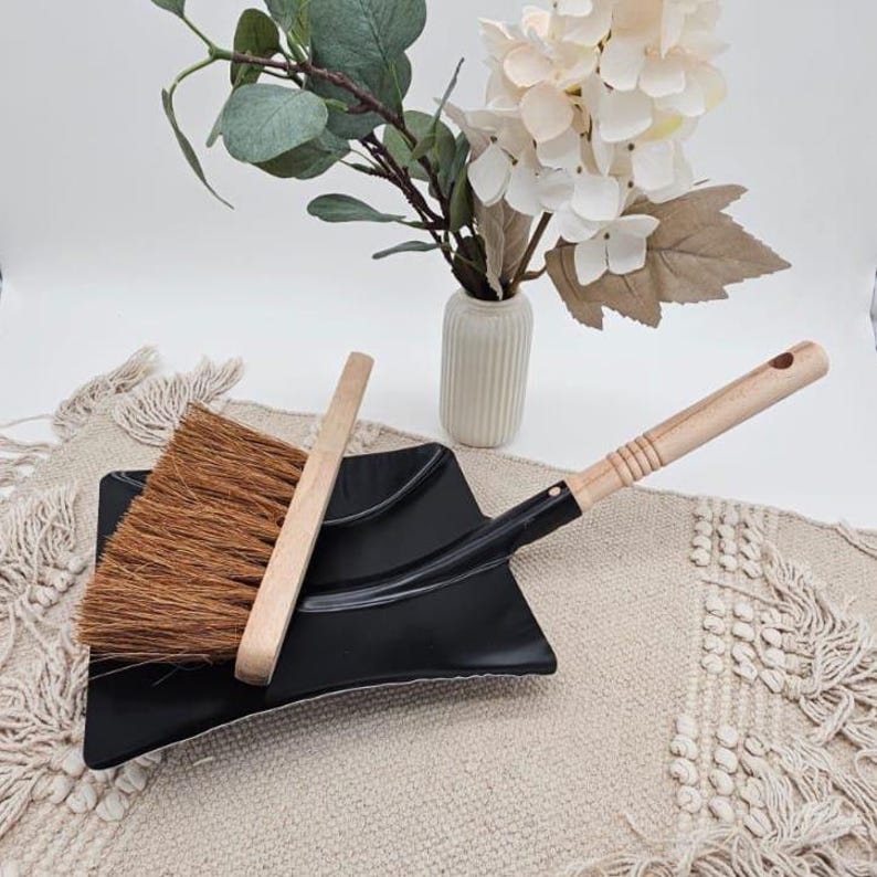 May include: A black dustpan with a wooden handle and a small broom with a wooden handle. The broom has brown bristles. The dustpan and broom are on a beige surface with a vase of flowers in the background.