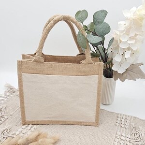 May include: A beige burlap tote bag with a cream-colored front pocket and rope handles. The bag is sitting on a textured beige surface. A vase of white flowers and greenery is in the background.