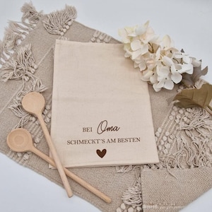 Könnte beinhalten: Beige Stoffbeutel mit dem Text "Bei Oma schmeckt's am besten" und einem Herzsymbol. Zwei Holzlöffel liegen auf einem beigen gewebten Platzset. Weiße Blumen sind auf der rechten Seite des Beutels angeordnet.