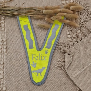 May include: A yellow safety vest with reflective strips and a dinosaur design. The vest has the name "Felix" printed on it.