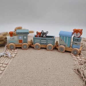 Könnte beinhalten: Ein hölzerner Spielzeugzug mit einer blauen und orangenen Lokomotive, einem blauen und grauen Güterwagen und einem blauen und weißen Schlusslicht. Die Lokomotive trägt den Text "Till" und "07.11.2023". Der Güterwagen trägt den Text "4200g" und "12:43 Uhr". Das Schlusslicht trägt den Text "54 cm". Der Zug ist mit Fuchs- und Hirschfiguren verziert.