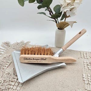 May include: A wooden dustpan and brush set with natural bristles. The brush handle is light-colored wood, and the dustpan is silver. The brush has the text "Scherben bringen Gl&uuml;ck! KERSTIN & OLLI" engraved on it. The set is on a beige surface.