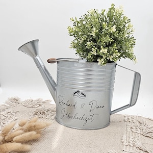 Könnte beinhalten: Eine verzinkte Metall-Gießkanne mit Griff und Ausguss, gefüllt mit einer grün-weißen Pflanze. Die Kanne hat ein dekoratives Design mit dem Text "Reinhard & Diana Silberhochzeit" und zwei Eheringen.