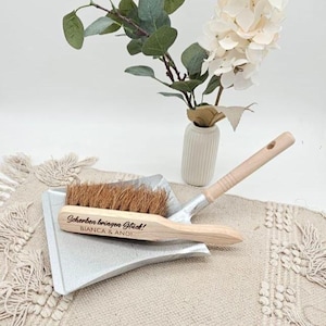 May include: A small, silver dustpan with a wooden-handled brush. The brush has brown bristles and the text "Scherben bringen Gl&uuml;ck!" and "BIANCA & ANDI" is engraved on the wooden handle. A vase with white flowers and greenery is in the background.