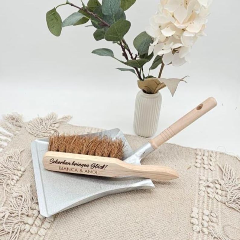 May include: A small, silver dustpan with a wooden-handled brush. The brush has brown bristles and the text "Scherben bringen Gl&uuml;ck!" and "BIANCA & ANDI" is engraved on the wooden handle. A vase with white flowers and greenery is in the background.