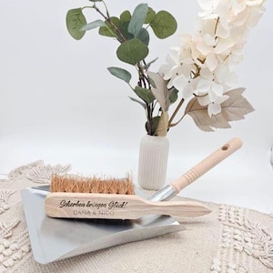 May include: A wooden dustpan and brush set with the text "Scherben bringen Gl&uuml;ck! DANA & NICO" engraved on the brush. The brush has natural bristles and a light wood handle. A small vase with flowers and greenery is in the background.