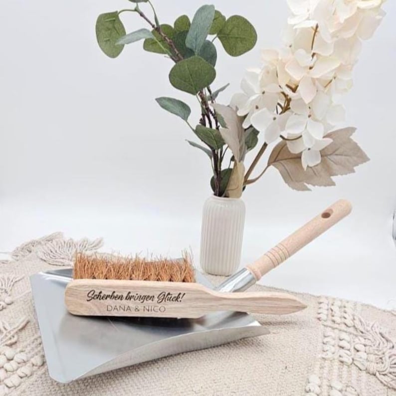 May include: A wooden dustpan and brush set with the text "Scherben bringen Gl&uuml;ck! DANA & NICO" engraved on the brush. The brush has natural bristles and a light wood handle. A small vase with flowers and greenery is in the background.
