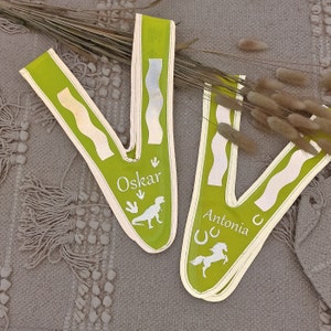 May include: Two green fabric sashes with white text and images. The sash on the left says "Oskar" and has a dinosaur silhouette. The sash on the right says "Antonia" and has a horse silhouette and a horseshoe.