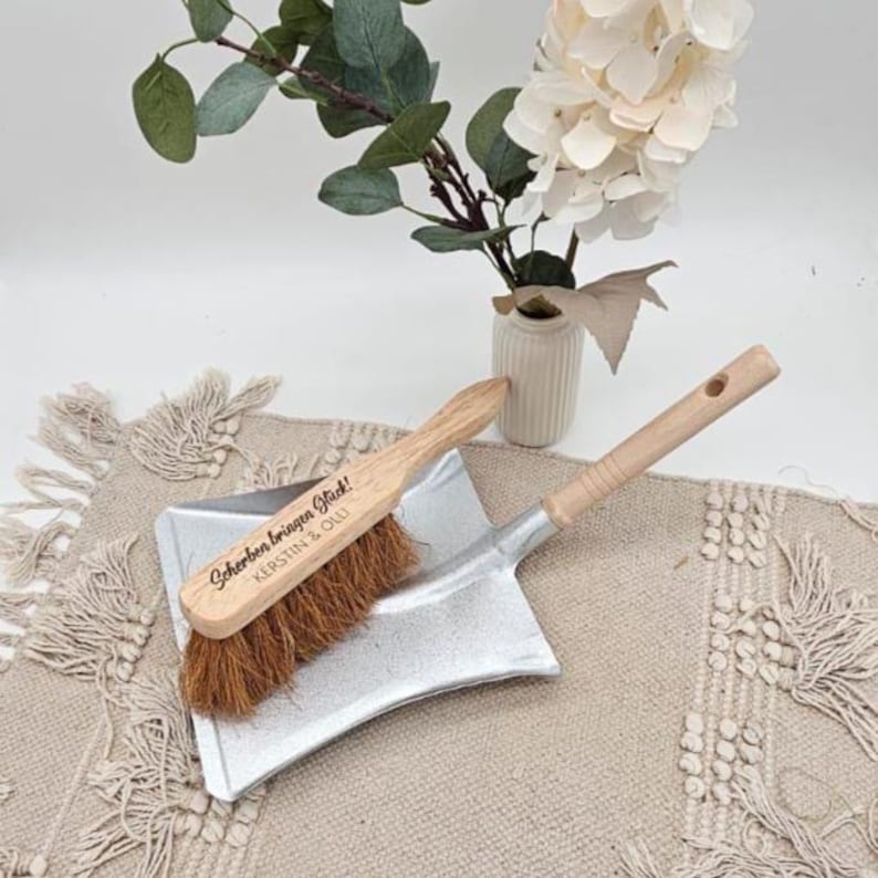 May include: A small, silver dustpan with a wooden handle and a matching brush. The brush has a wooden handle and brown bristles. The text "Scherben bringen Gl&uuml;ck!" and "KERSTIN & OLLI" is printed on the brush. The items are on a beige woven mat.