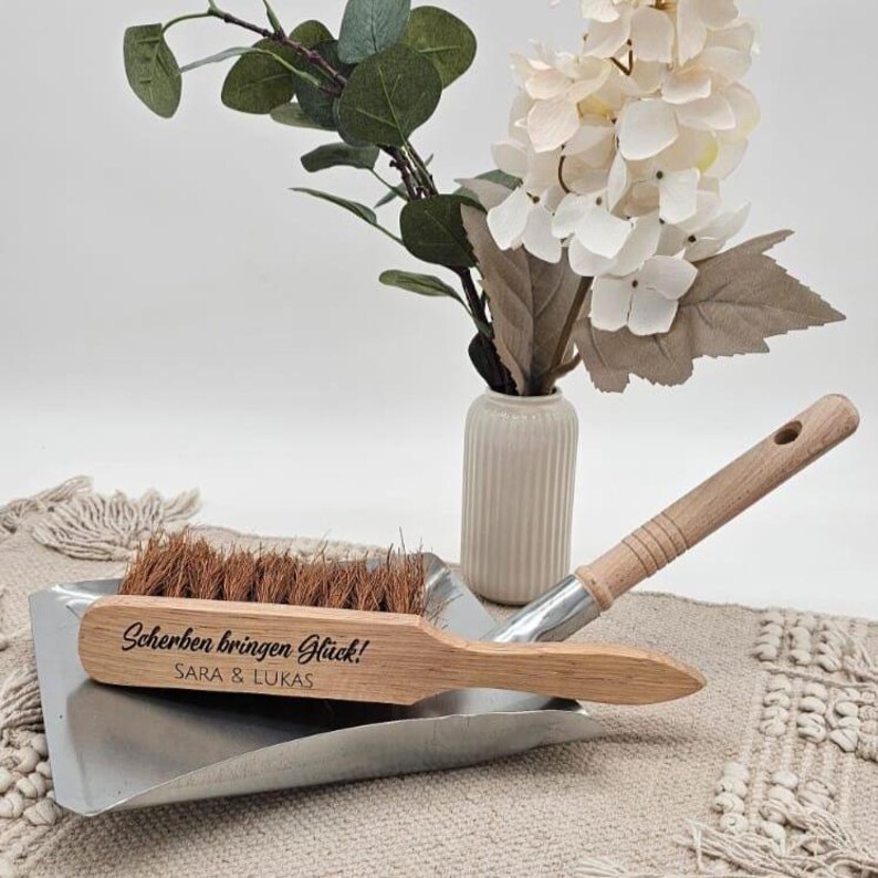 May include: A wooden dustpan and brush set with the text "Scherben bringen Gl&uuml;ck!" and the names "SARA & LUKAS" engraved on the brush. The brush has brown bristles and a wooden handle. The set is on a metal dustpan.