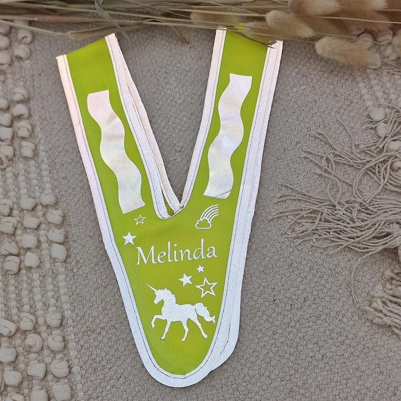 May include: A lime green safety vest with reflective strips and a white unicorn design. The vest has the name "Melinda" printed on it.
