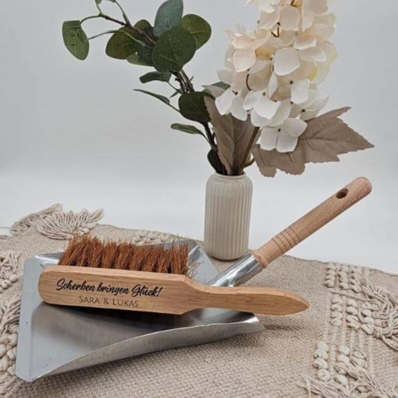 May include: A wooden dustpan and brush set with the text "Scherben bringen Gl&uuml;ck!" and the names "SARA & LUKAS" engraved on the brush. The brush has brown bristles and a wooden handle. The dustpan is made of silver metal. A vase of white flowers is in the background.