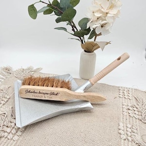 May include: A wooden and metal dustpan set with a natural fiber brush. The brush handle has the text "Scherben bringen Gl&uuml;ck! Franzi & Tim". A small white vase with flowers is in the background.