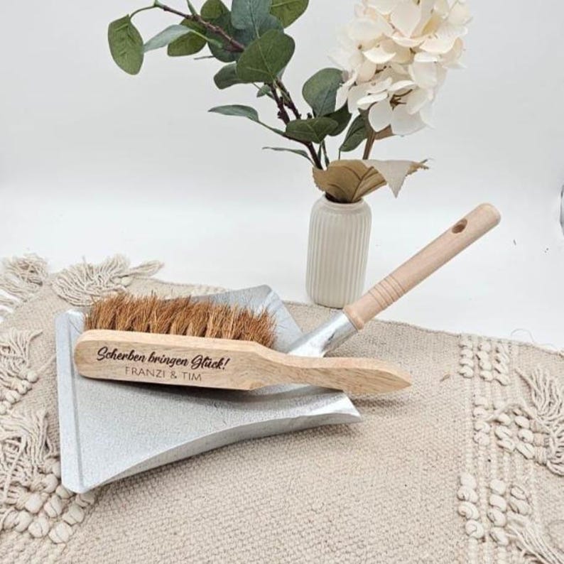 May include: A wooden and metal dustpan set with a natural fiber brush. The brush handle has the text "Scherben bringen Gl&uuml;ck! Franzi & Tim". A small white vase with flowers is in the background.