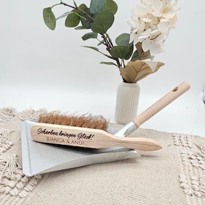 May include: A dustpan and brush set with the text "Scherben bringen Gl&uuml;ck!" and "BIANCA & ANDI" engraved on the wooden brush. The set is silver and natural wood, with a light brown brush. A vase with flowers is in the background.