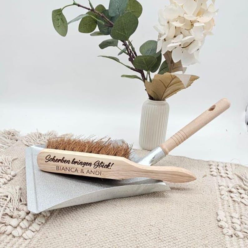 May include: A dustpan and brush set with the text "Scherben bringen Gl&uuml;ck!" and "BIANCA & ANDI" engraved on the wooden brush. The set is silver and natural wood, with a light brown brush. A vase with flowers is in the background.