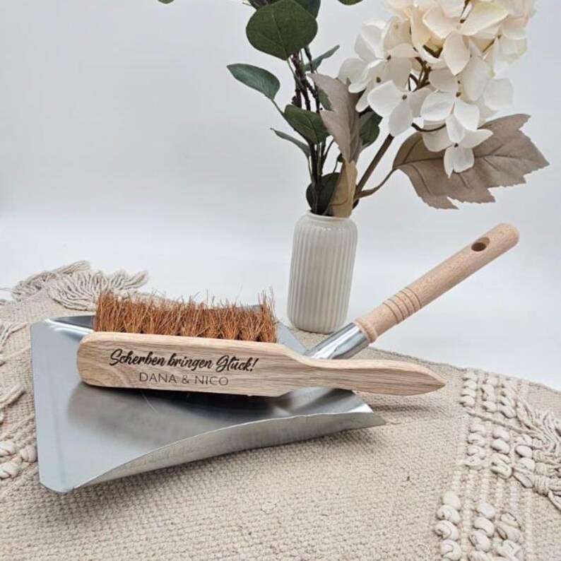 May include: A dustpan and brush set with a wooden handle and a metal dustpan. The brush has brown bristles. The wooden handle of the brush has the text "Scherben bringen Gl&uuml;ck!" and "DANA & NICO". A vase with flowers is in the background.
