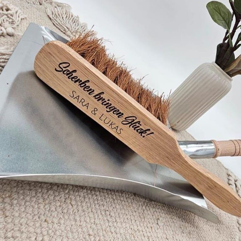 May include: A wooden dustpan and brush set. The brush has a light brown wooden handle with the text "Scherben bringen Gl&uuml;ck!" and the names "SARA & LUKAS" printed on it. The brush bristles are brown. The dustpan is silver.
