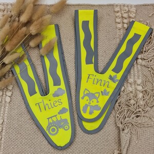 May include: Two yellow safety vests with reflective strips. One vest has the name "Thies" and a tractor design. The other vest has the name "Finn" and a fox design.