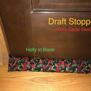 May include: A long, cylindrical draft stopper positioned at the base of a dark brown wooden door. The fabric features a black background with a repeating pattern of green holly leaves and red berries. Text reads Draft Stopper (100% Cedar filled).