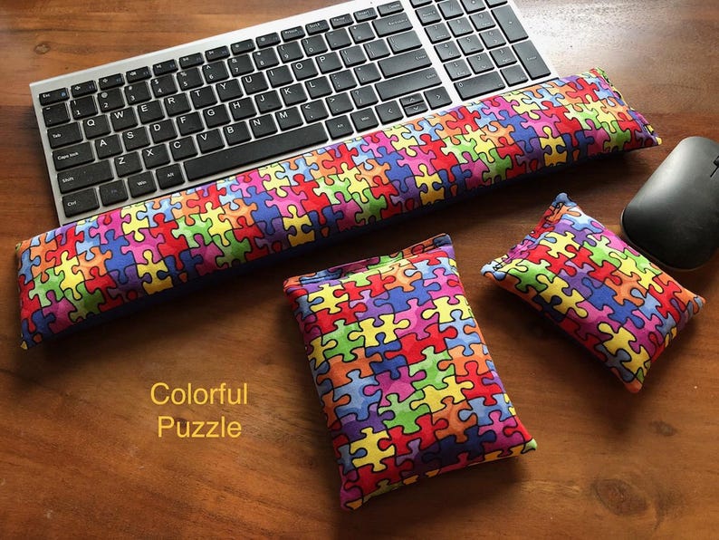 May include: A keyboard, mouse, and three colorful puzzle-patterned cushions rest on a wooden surface. The cushions are in various sizes, with a long one for the wrist and two smaller ones. The text "Colorful Puzzle" is visible.