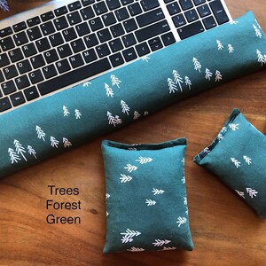 May include: A forest green keyboard wrist rest and two matching square cushions. The fabric features a white tree pattern. The items are displayed on a wooden surface next to a computer keyboard and mouse.