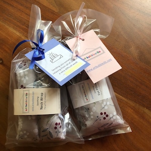 Pu&ograve; includere: Due confezioni regalo in sacchetti di cellophane trasparente, ciascuna con un'etichetta e un nastro. Un set contiene un "Soothing Neck Wrap" e l'altro un "Eye Pillow". Il tessuto ha uno sfondo grigio con fiori bianchi e bacche rosse. Le etichette riportano "artbubblesllc.com".