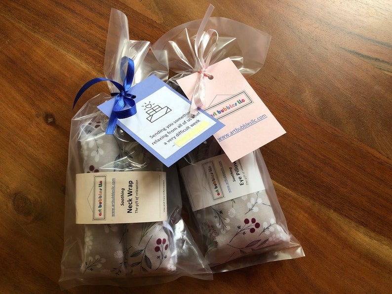 Pu&ograve; includere: Due confezioni regalo in sacchetti di cellophane trasparente, ciascuna con un'etichetta e un nastro. Un set contiene un "Soothing Neck Wrap" e l'altro un "Eye Pillow". Il tessuto ha uno sfondo grigio con fiori bianchi e bacche rosse. Le etichette riportano "artbubblesllc.com".