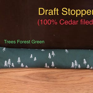 May include: A forest green draft stopper with a white tree pattern. The text "Draft Stopper (100% Cedar filed)" is in yellow and red. The text "Trees Forest Green" is in green.