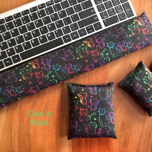 May include: Black keyboard wrist rest, mouse pad, and small pillow set. The fabric features a repeating pattern of colorful cat illustrations. The text "Cats in Black" is visible on the wooden surface.