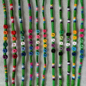 May include: A selection of colourful friendship bracelets. Each bracelet has a green woven base with flower-shaped designs in pink, yellow, blue, and white. The bracelets are displayed vertically, showing their intricate patterns.