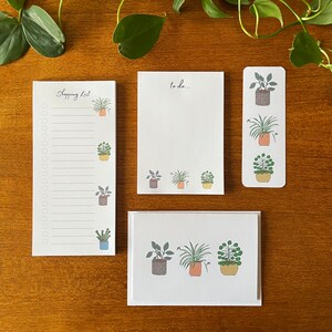 May include: Set of four white notepads with a plant theme. Each notepad has a different design featuring potted plants with green leaves and brown pots. The notepads have the words "Shopping List" and "to do..." printed on them.